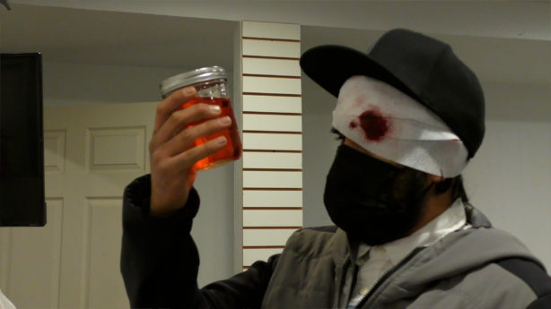 A person with a masked and bandaged face looks into a jar filled with an amber-colored substance.