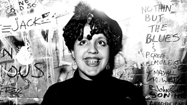 The woman of color grinning & showing off her braces in front of a heavy graffitied wall is Poly Styrene! 
