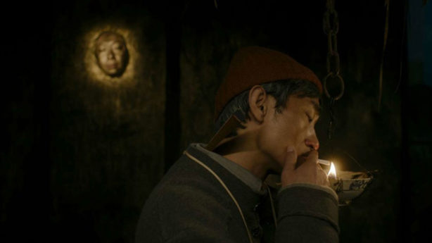 A man is smoking in a dark room with a sining token on the wall at the back.