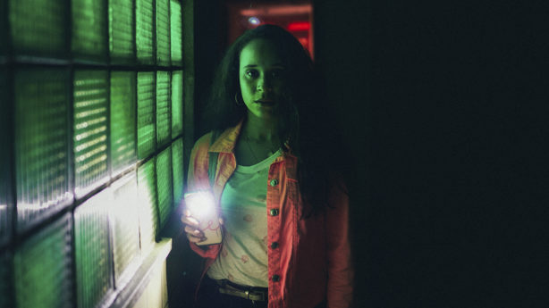 A woman, looking scared, is alone in a dark room with her flashlight on.